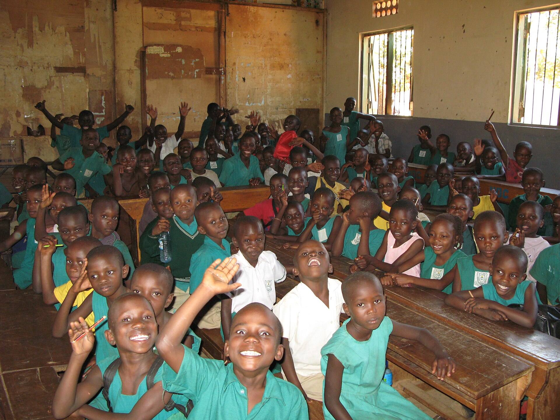 A packed primary classroom of children in green uniforms, many with their hands raised